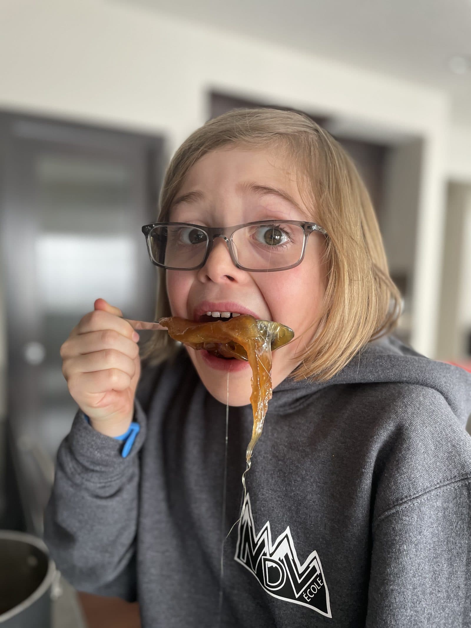 Kid eating homemade tire d'erable maple taffy off a stick, eyes wide, sticky strands of cooled syrup running down