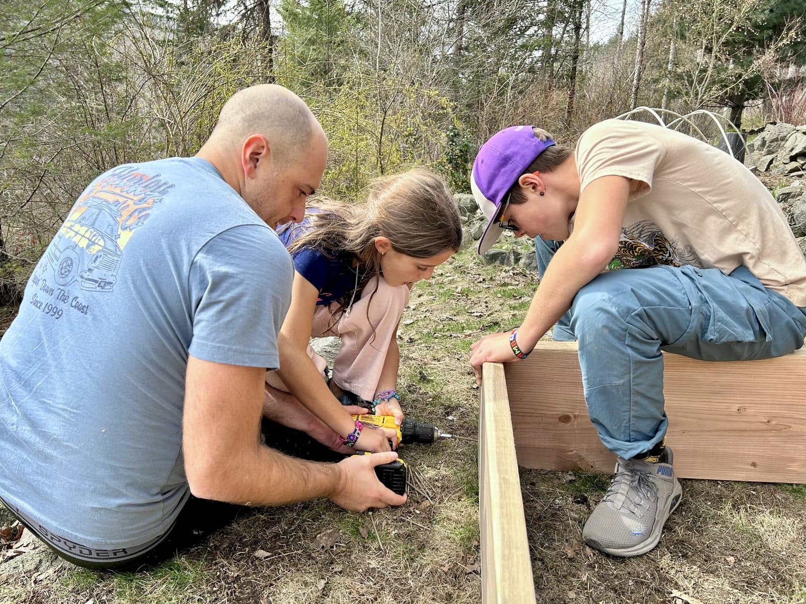 Dad and two kids building a wooden garden bed together with a drill and lumber, real-world engineering happening in the backyard