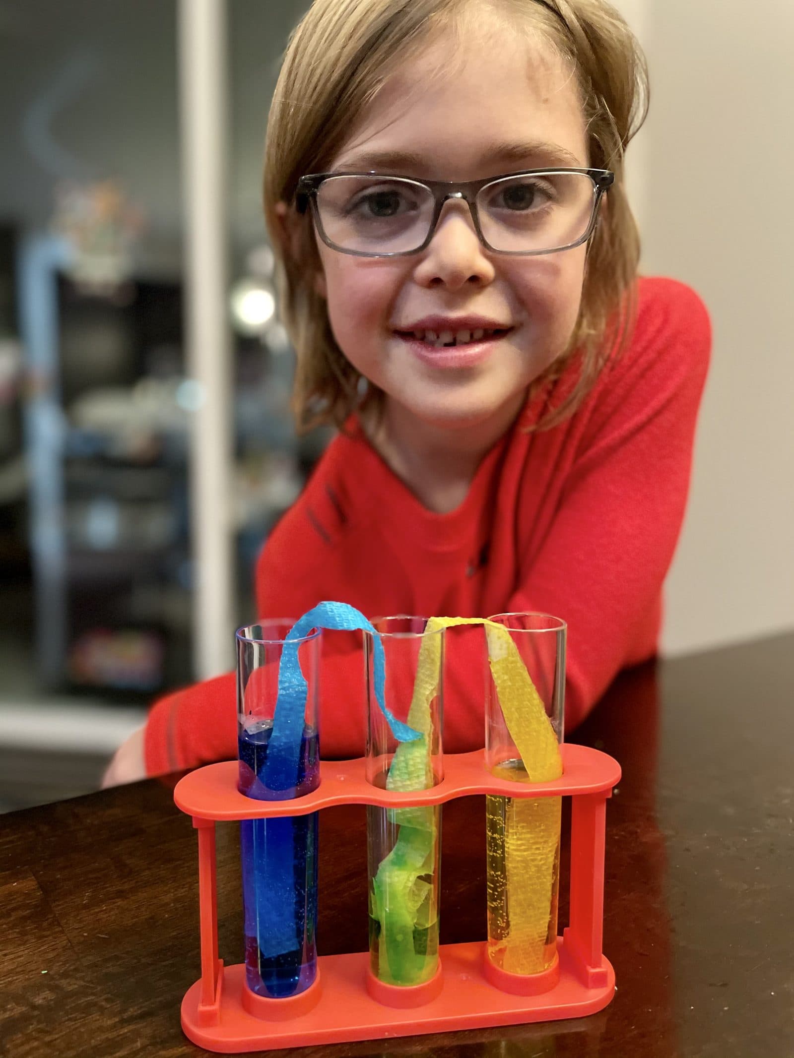 Young kid standing behind a tray of test tubes filled with blue, green, and yellow liquid, beaming about a homemade chemistry experiment