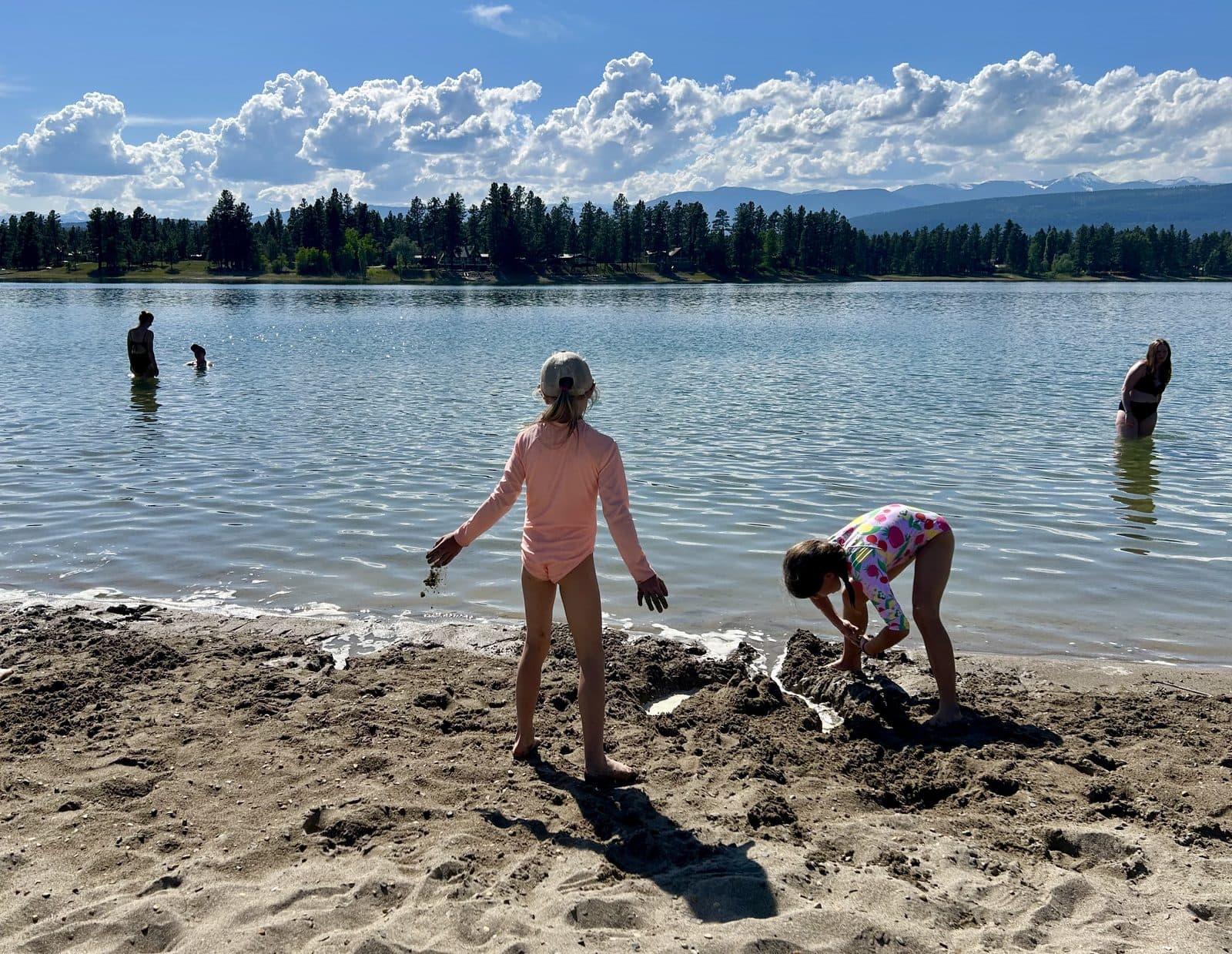 Two kids actively shaping sand at the edge of a mountain lake on a summer afternoon, the kind of hands-on outdoor STEM that makes summer count without a camp