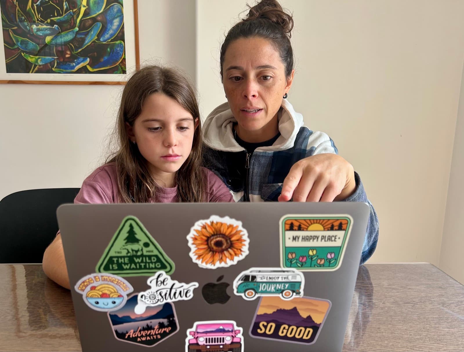 Child typing on a laptop with a thoughtful expression, parent watching nearby