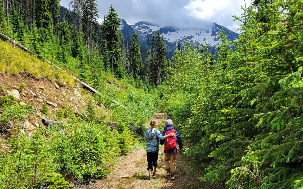 Two kids walking a mountain trail together, one with an arm around the other in a moment of care and support, with a snowy peak behind them