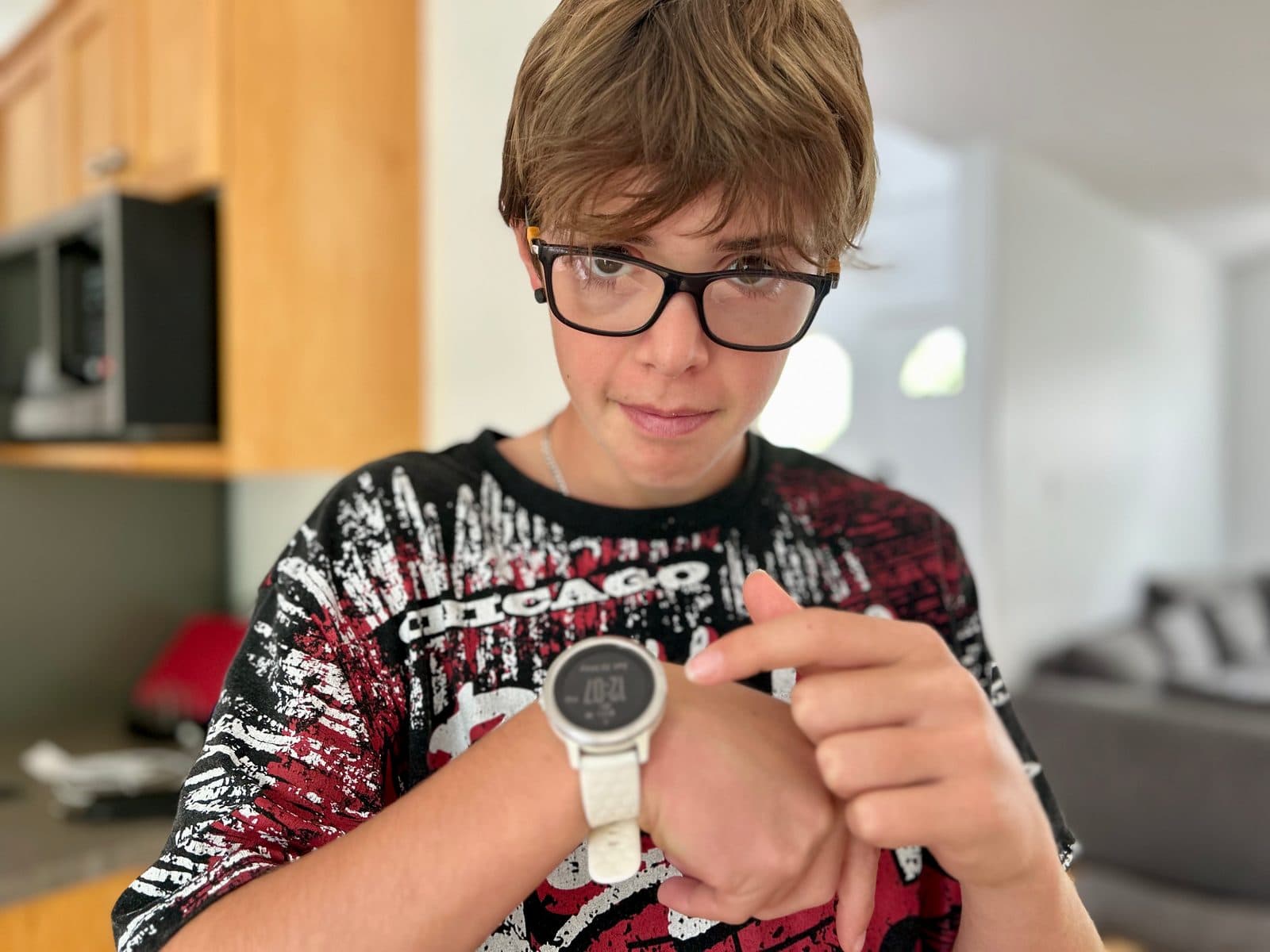 A pre-teen boy in glasses pointing at the time on his wristwatch in the kitchen, focused expression