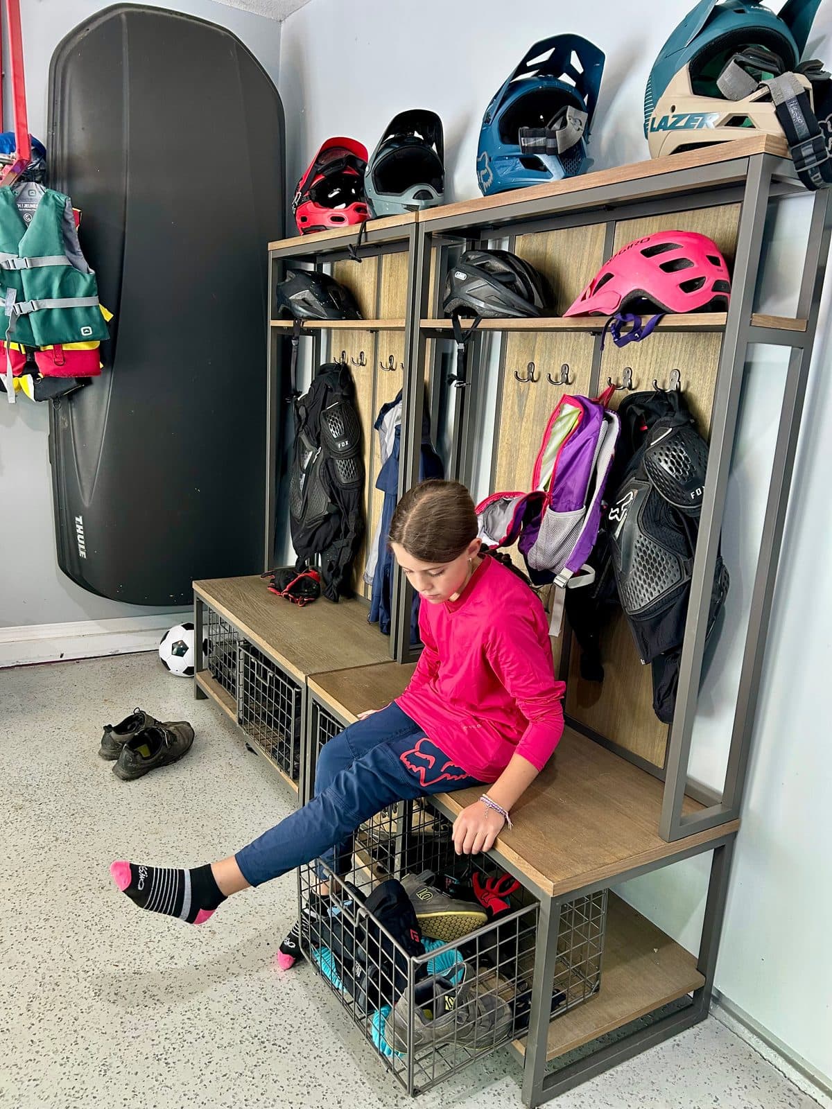 A kid putting on socks and shoes for a bike ride, helmets and gear hanging on the wall behind her