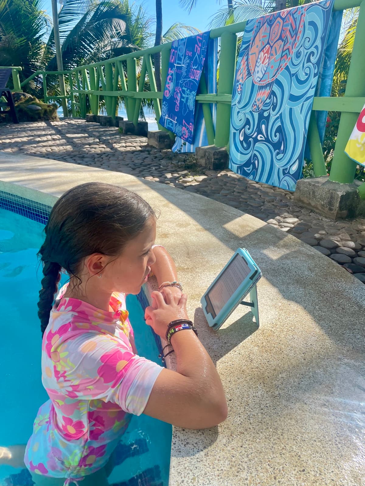 My daughter reading on her tablet by the pool with colourful towels hanging on the railing behind her