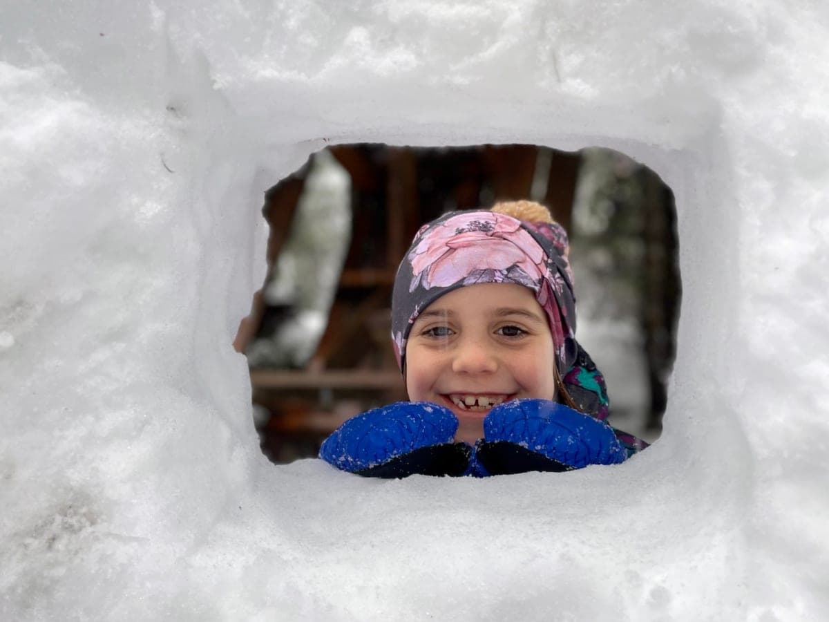 A smiling girl in a flowered hat and blue mittens peeking through a window cut into a thick snow wall
