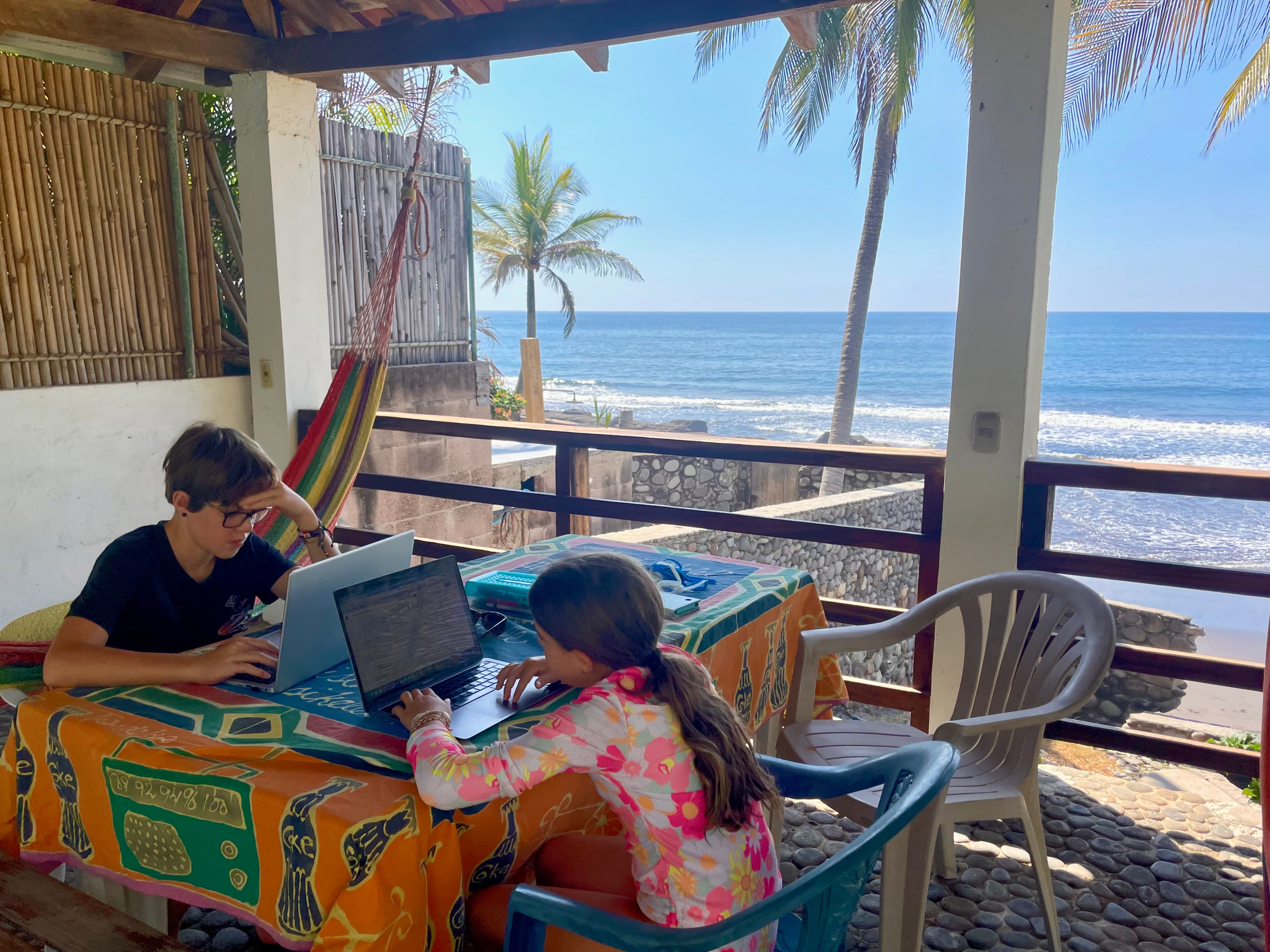 Two kids working on laptops on a patio overlooking the ocean and palm trees